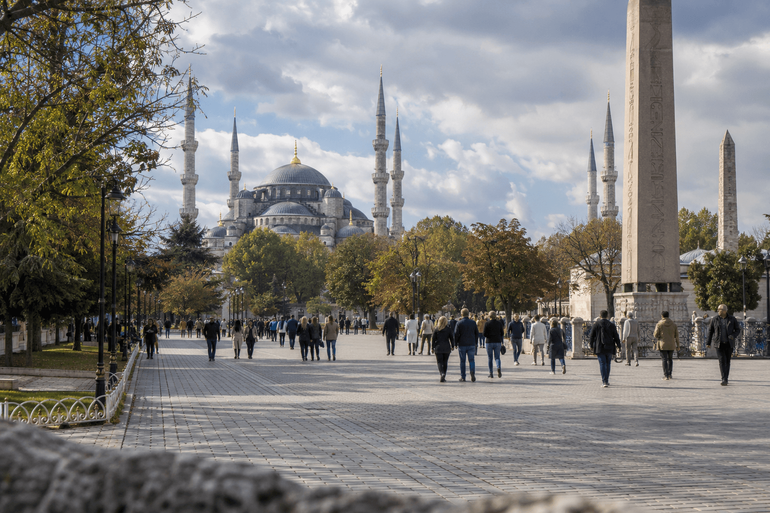 Visitors in light jackets walking the broad paved approach to the Blue Mosque past the Hippodrome obelisk