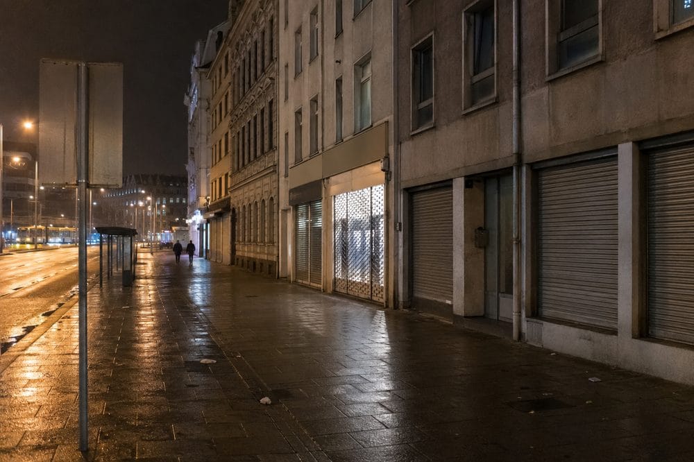 A worn street on the fringe of a station district at dusk, roller shutters down and wet pavement reflecting shopfront light