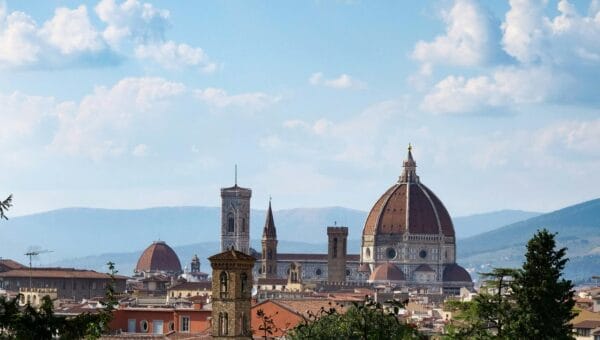 Florence rooftops with Brunelleschi's dome and Giotto's flat-topped campanile seen from a garden terrace, cypress branches framing the left of the view