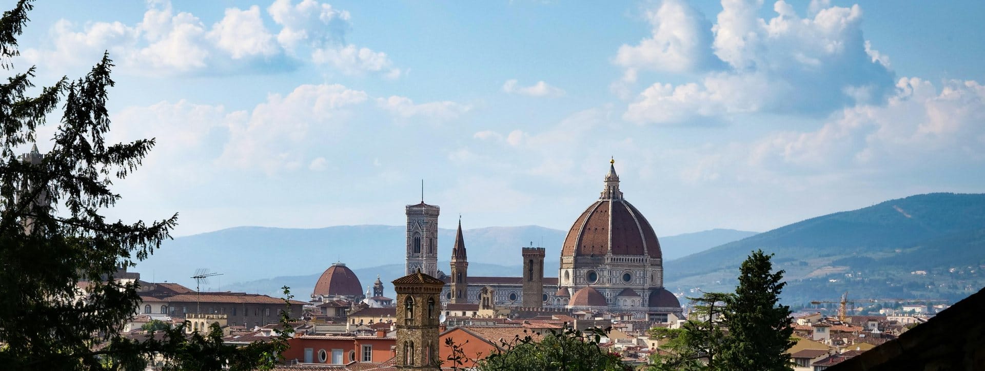 Florence rooftops with Brunelleschi's dome and Giotto's flat-topped campanile seen from a garden terrace, cypress branches framing the left of the view