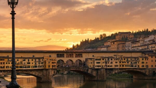 The Ponte Vecchio at sunset seen from the Arno embankment, its three arches and the Vasari Corridor windows reflected in still water