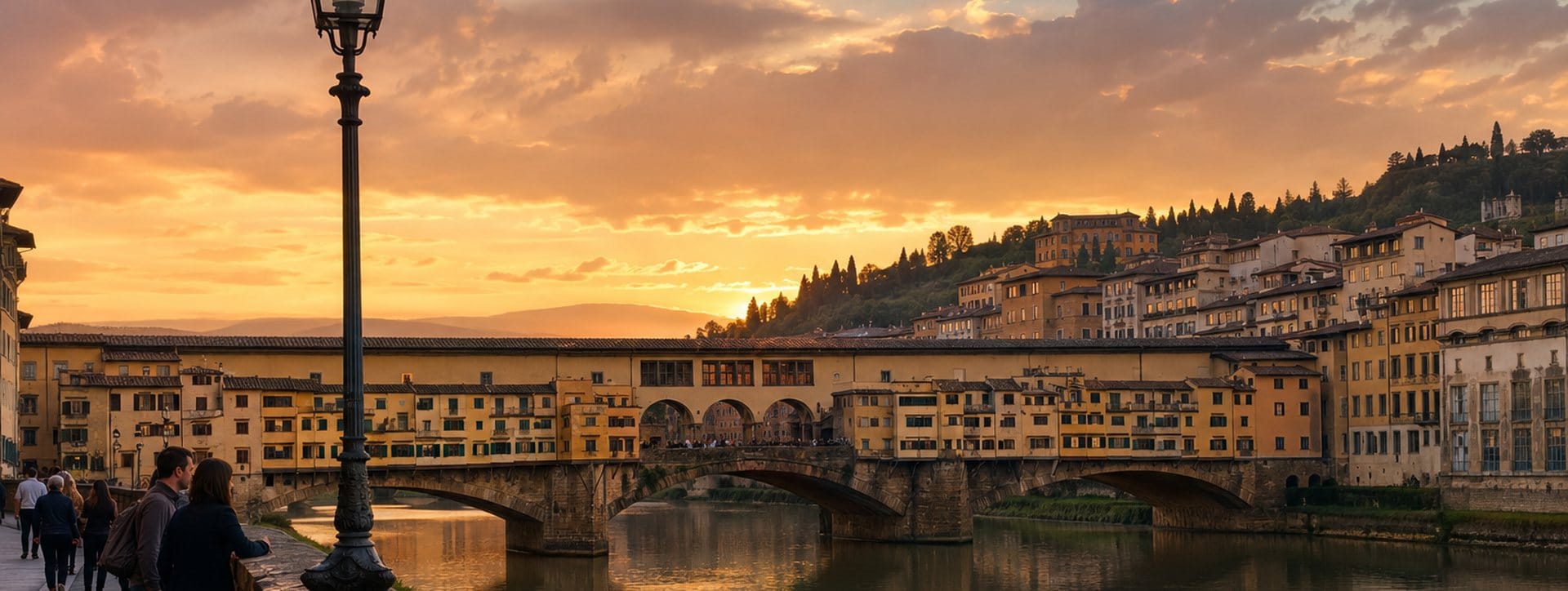 The Ponte Vecchio at sunset seen from the Arno embankment, its three arches and the Vasari Corridor windows reflected in still water