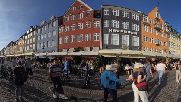 Crowds walking past the cafe tables and colorful townhouses along the Nyhavn canal in Copenhagen on a sunny day