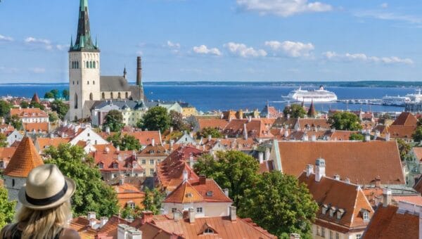Two travelers look out from a stone wall over Tallinn Old Town's red roofs toward St Olaf's Church spire and a harbor with a white cruise ship.