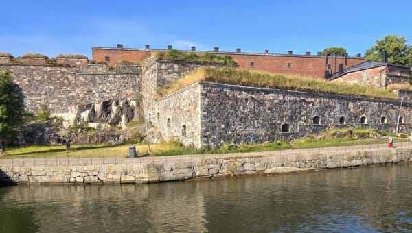 Stone fortress walls and red-brick barracks of Suomenlinna above the water on a sunny morning, with a few people walking the waterfront path